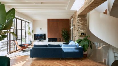 modern living room with blue sofa, spiral staircase, herringbone floors, a potted plant in the foreground, and two by the TV, with an armchair near the floor-to-ceiling windows