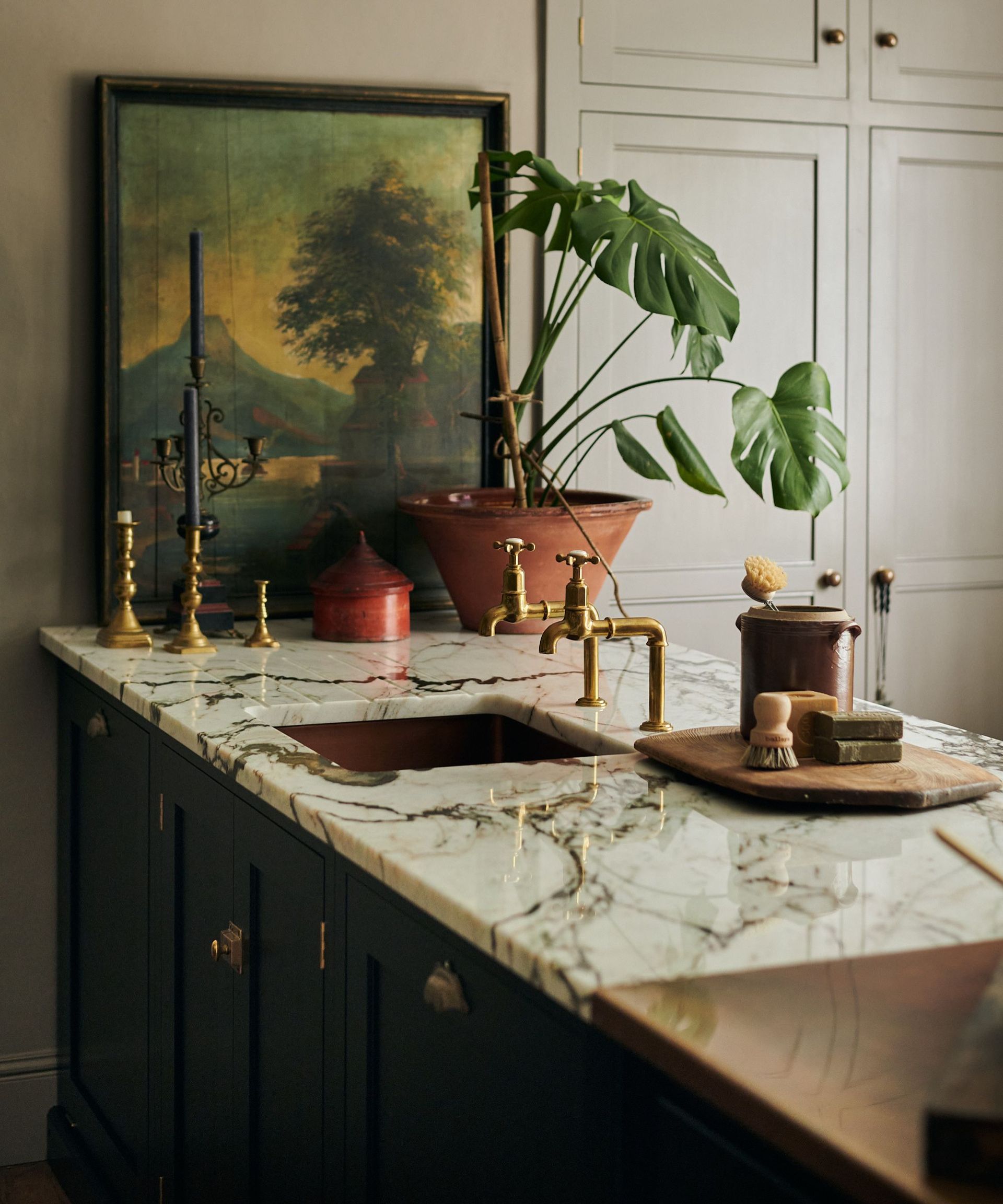 Marble kitchen island with dark gray veins, with copper sink and brass faucets. There is a natural wooden bristled cleaning brush and sponges on a wooden board.