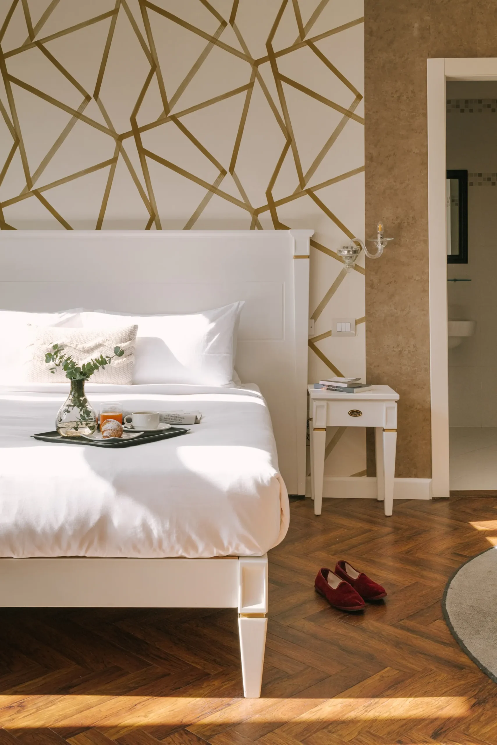 A sunlit hotel run features wooden floor, textural walls with a geometric, gold-on-white motif, white bedding on which a breakfast tray rests, and matching white furniture in wood.