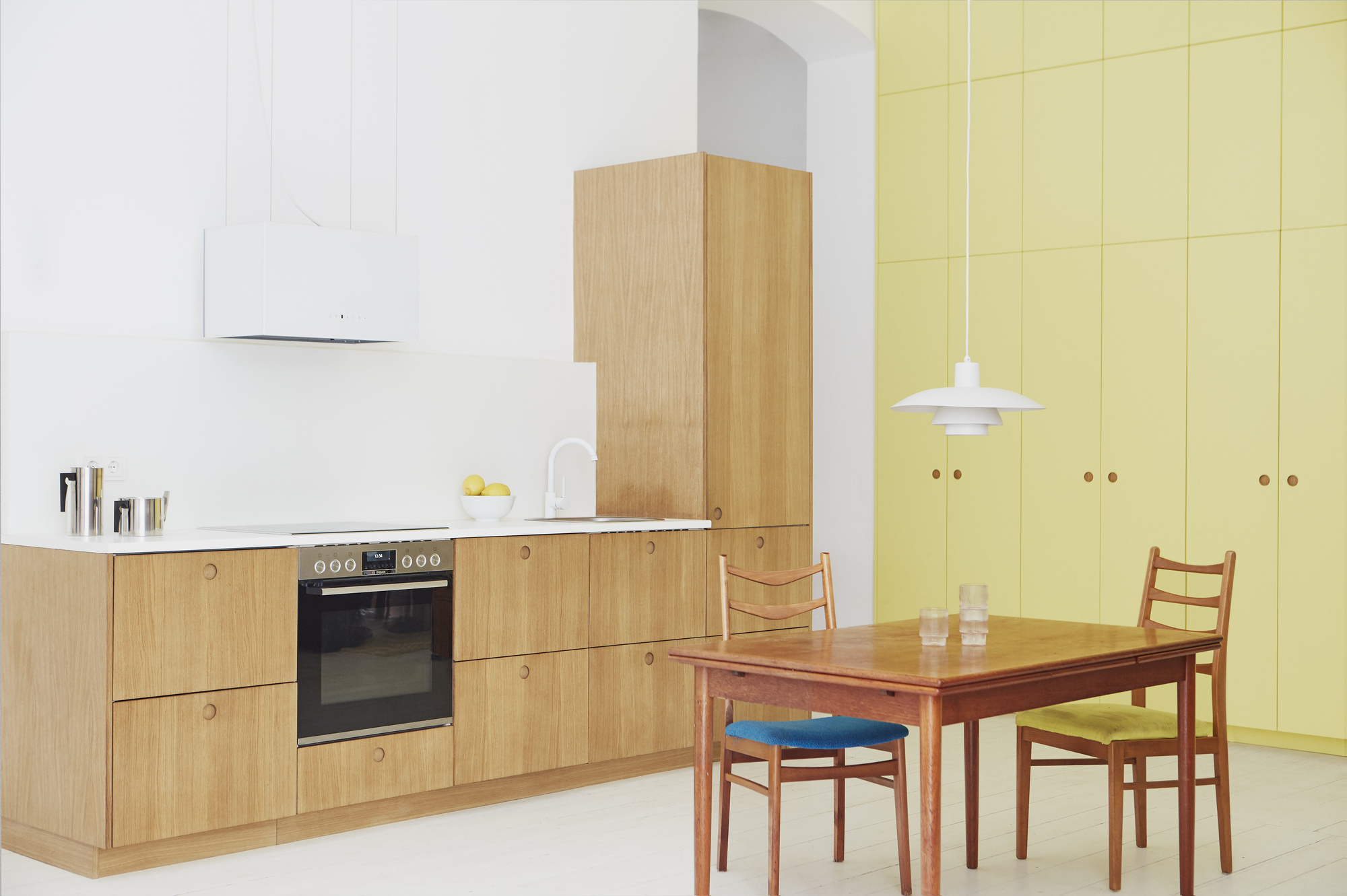 yellow kitchen cabinets, timber kitchen with white countertops, white tap, white walls, white painted floorboards, and timber dining table with timber chairs