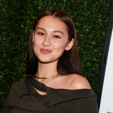 a woman with brown hair wearing an off-shoulder top posing in front of a grass wall