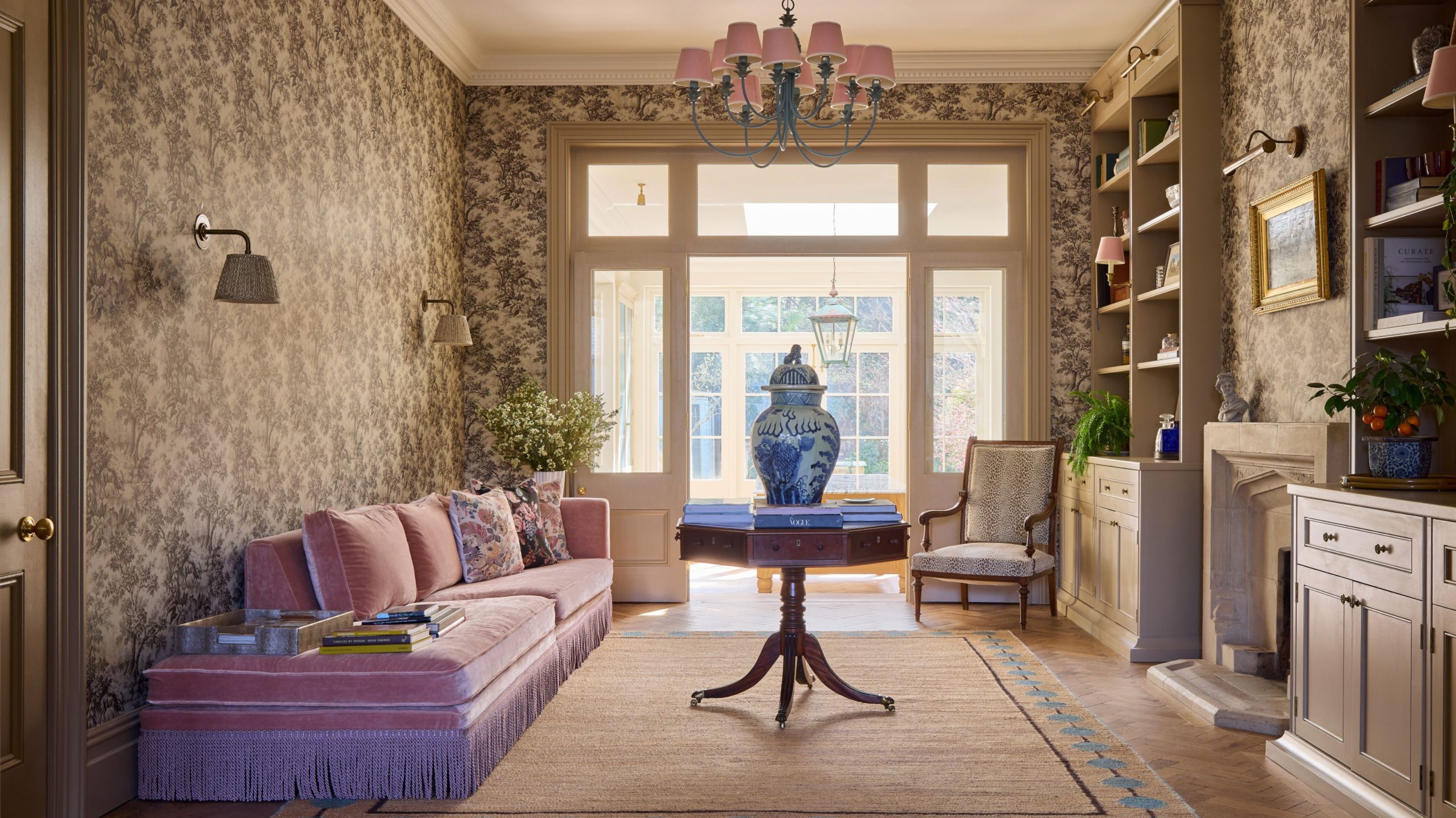 Elegant sitting room with damask wallpaper, a long pink velvet daybed with fringe, a central pedestal table with a large blue-and-white porcelain jar, and built-in shelving.