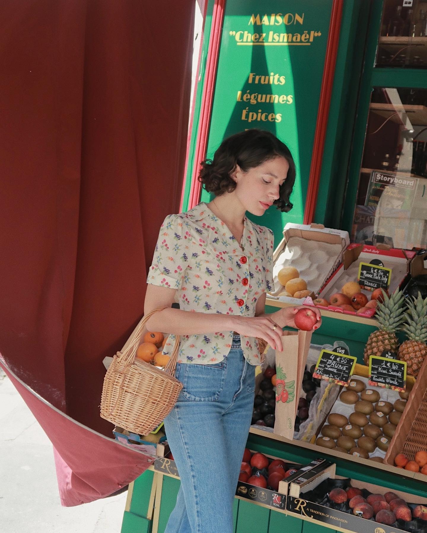Influencer @melodiebanfield stands in front of a fruit market wearing a floral blouse, patch-pocket jeans and a wicker basket.