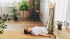 woman wearing white tshirt and khaki leggings on a pale orange yoga mat and wooden floor with her legs vertically up a wall. there are plants around her.