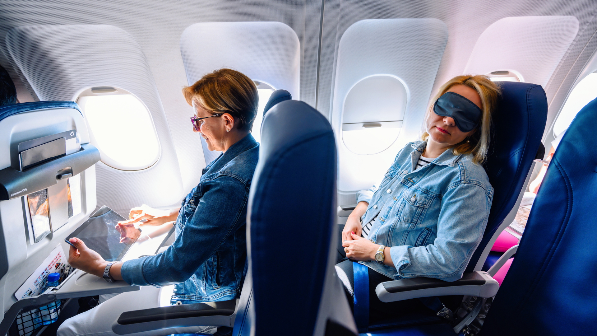 Two women sit in two rows of a plane - one looker at her tablet, one sleeping with an eye mask on