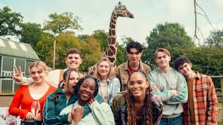 The Season 3 cast of Heartstopper smiling in front of the giraffe exhibit at a zoo 