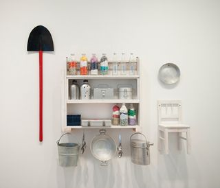 Left to right items displayed on a wall. A shovel, Three shelves with bottles and flasks on with three buckets underneath. A chair.