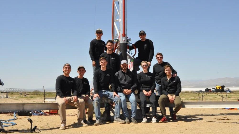 a small rocket with a group of people surrounding it, in a sandy area. blue sky is in behind