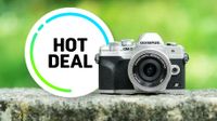 A silver and black Olympus OM-D E-M10 Mark IV camera sits on a stone ledge. Beside it, a circular graphic with "Hot Deal" in bold black text on a white background