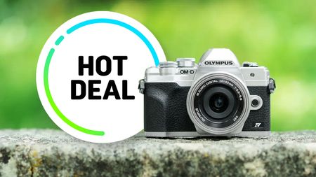 A silver and black Olympus OM-D E-M10 Mark IV camera sits on a stone ledge. Beside it, a circular graphic with "Hot Deal" in bold black text on a white background