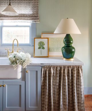 Kitchen with blue shaker cabinets, marble worktops, green table lamp and a fabric cabinet skirt in Penny Morrison fabric