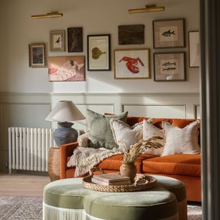 A living room with a gallery wall with picture lights and a table lamp on a side table next to the velvet orange sofa