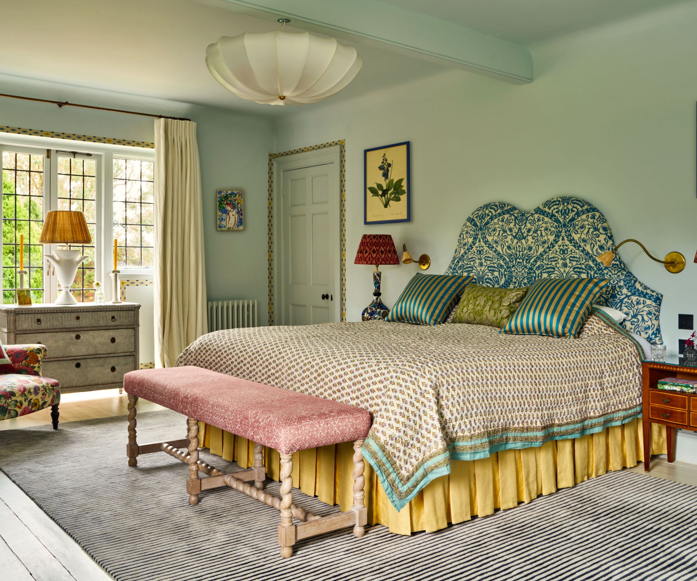 Bedroom painted in Farrow &amp;amp; Ball's Borrowed Light, a blue upholstered headboard, a yellow skirt, a pink bench, large windows with a dresser and lamp in front, and a floral armchair in the corner.