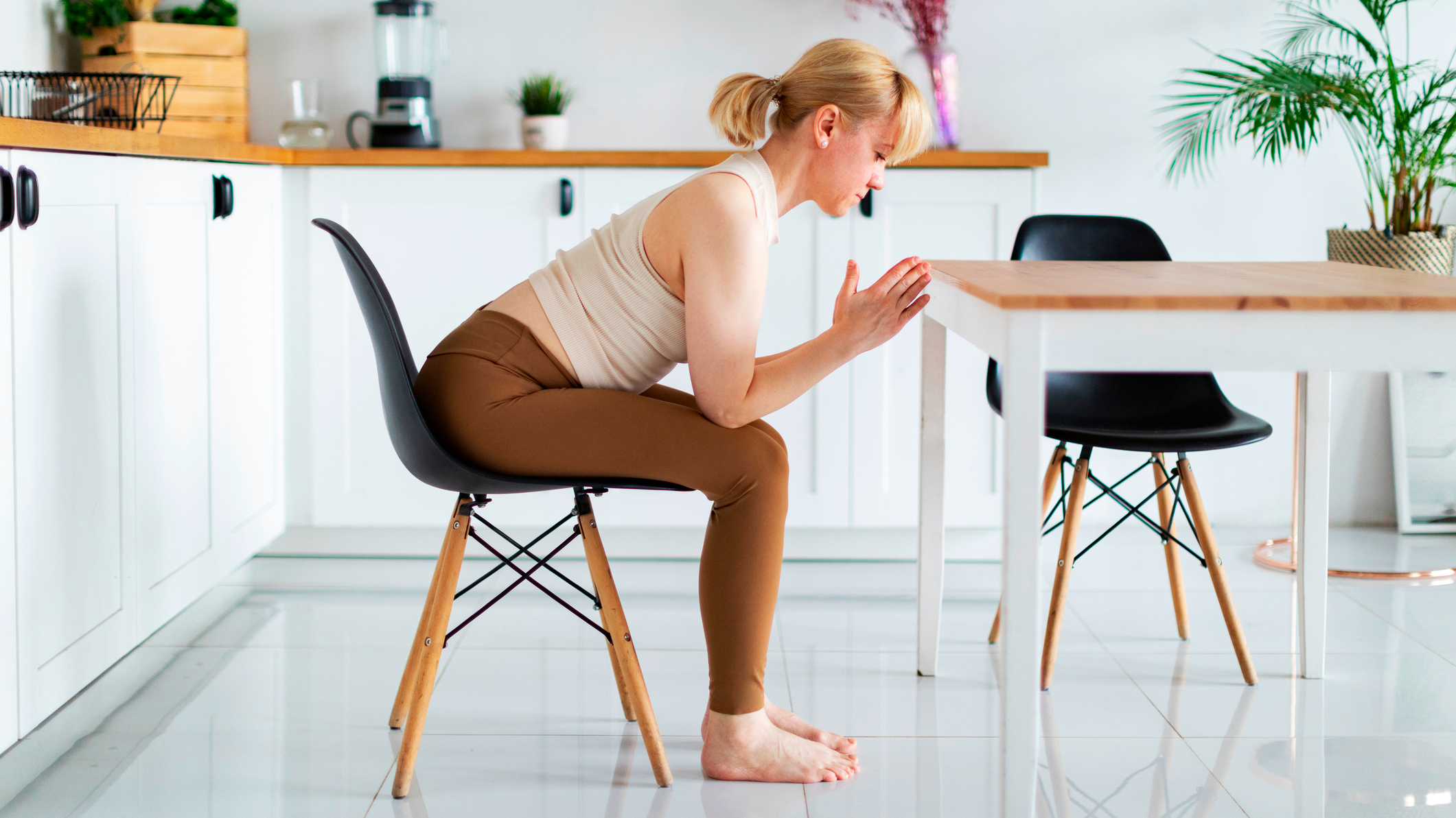 Woman wearing sportswear sits in chair in kitchen and leans forward from her hips