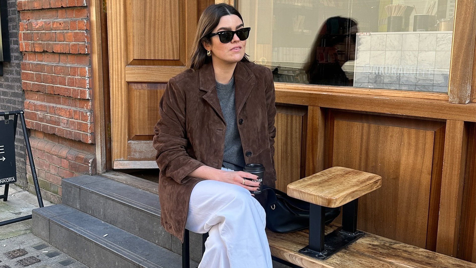British style influencer Anna Newton poses on a bench outside a cafe in London wearing Ray Ban wayfarer sunglasses, a brown suede blazer, a gray crewneck sweater, and white jeans