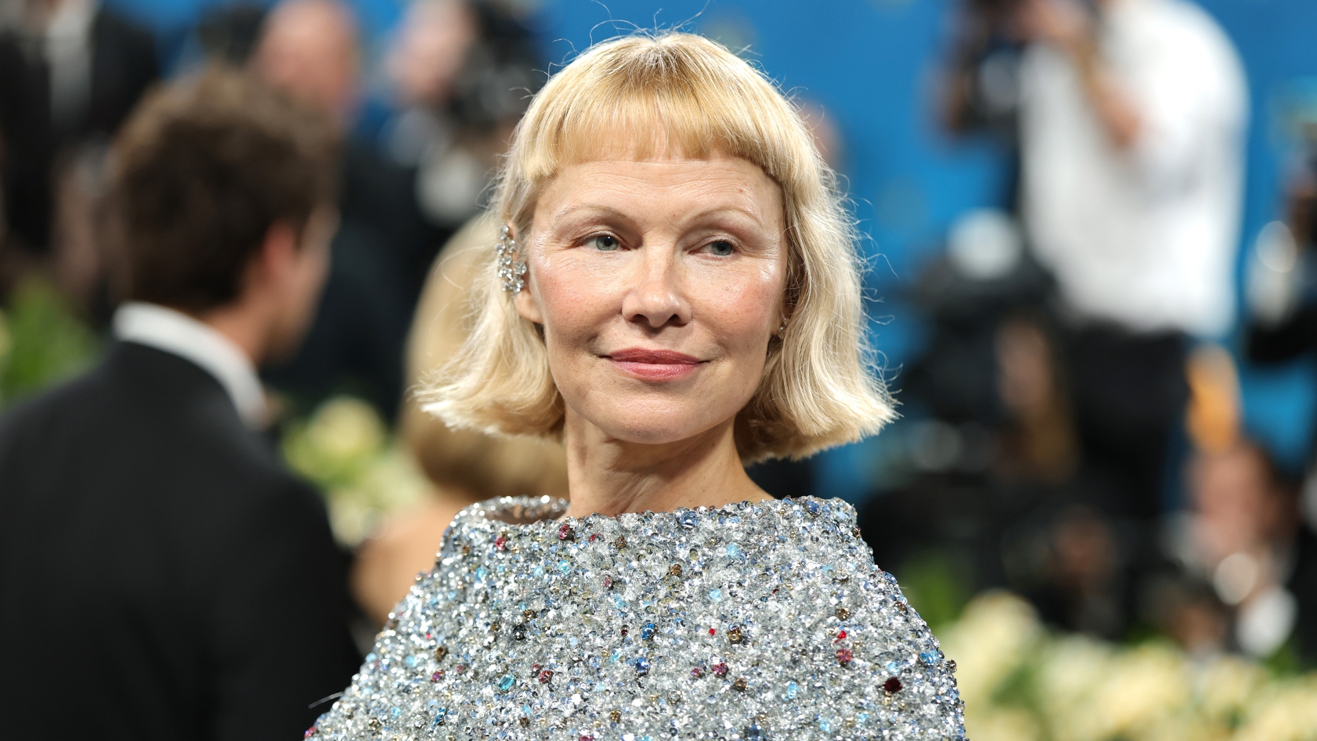 Image of Pamela Anderson smiling with short blonde jaw-length bob and baby bangs, wearing a silver jewel-embellished gown, at the 2025 Met Gala