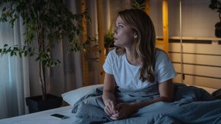 A woman sitting up in bed in the evening looking anxious.