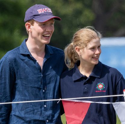 Lady Louise Windsor and Felix da Silva-Clamp wearing blue shirts and smiling outdoors in front of a rope fence with red and white bunting 