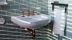 Sunlight coming through from the window in a stylish bathroom with green brick tiled wall and clean white grout. A white sink has gold taps and a white towel hangs from a rail