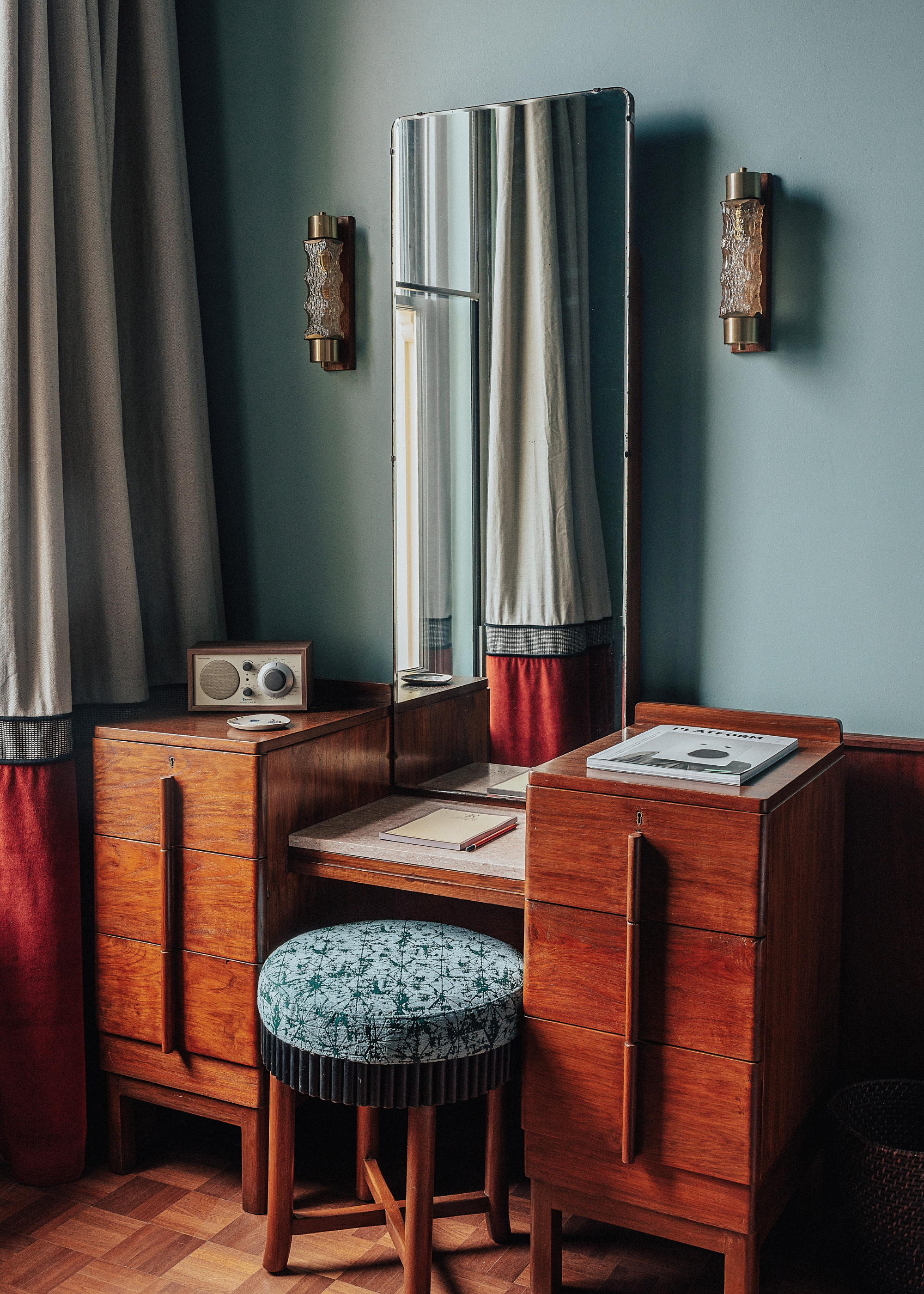 A vibrantly decorated hotel suite inspired by Bombay residences features buttery or tile blue walls adorned with original portraiture artworks, bespoke retro-fueled furnishings in wood, leather, and patterned fabric, and thriving plants.