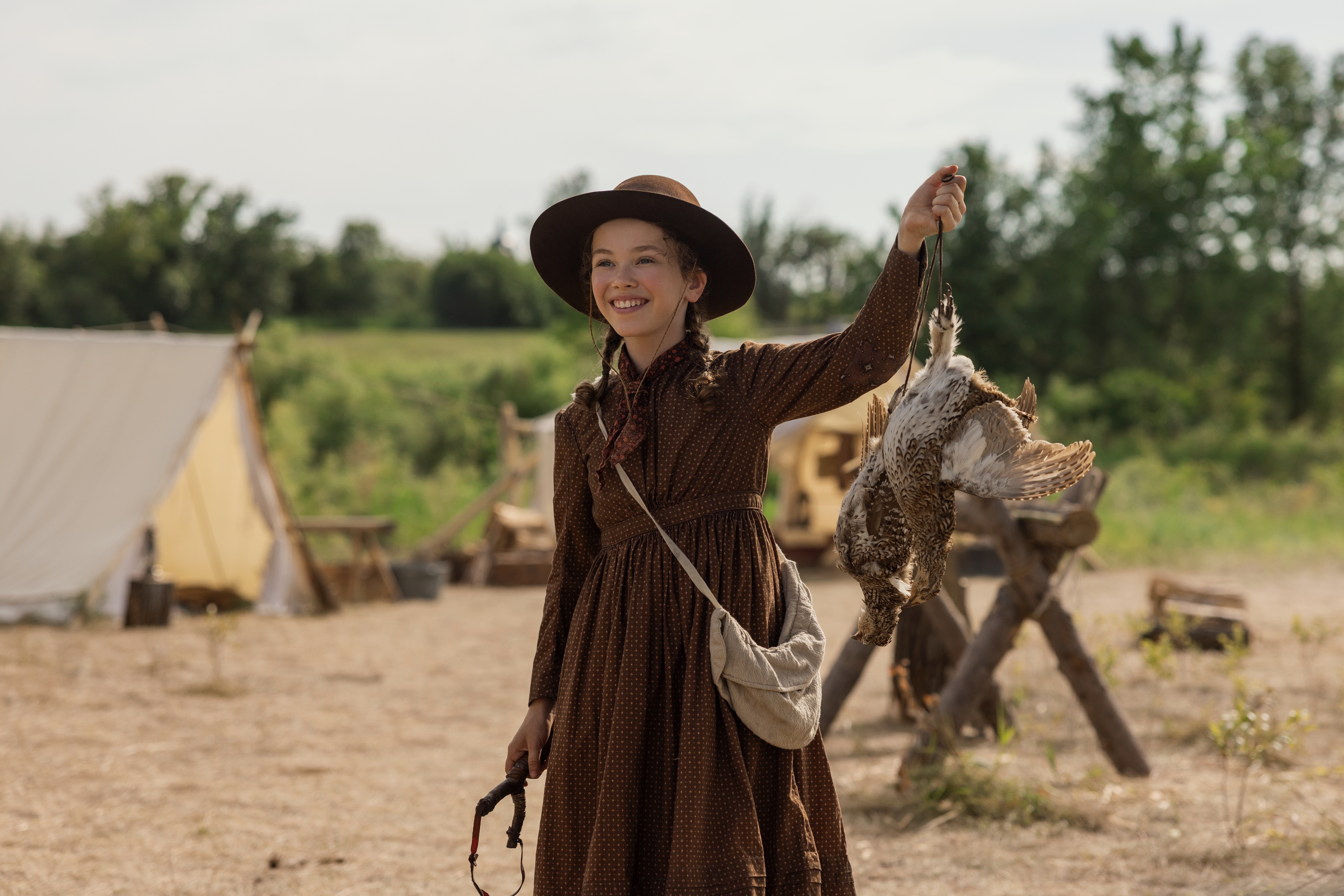 Actress Alice Halsey as Laura Ingalls waving while dressed in 19th century clothing and standing in front of a campsite, in Netflix's 'Little House on the Prairie.'