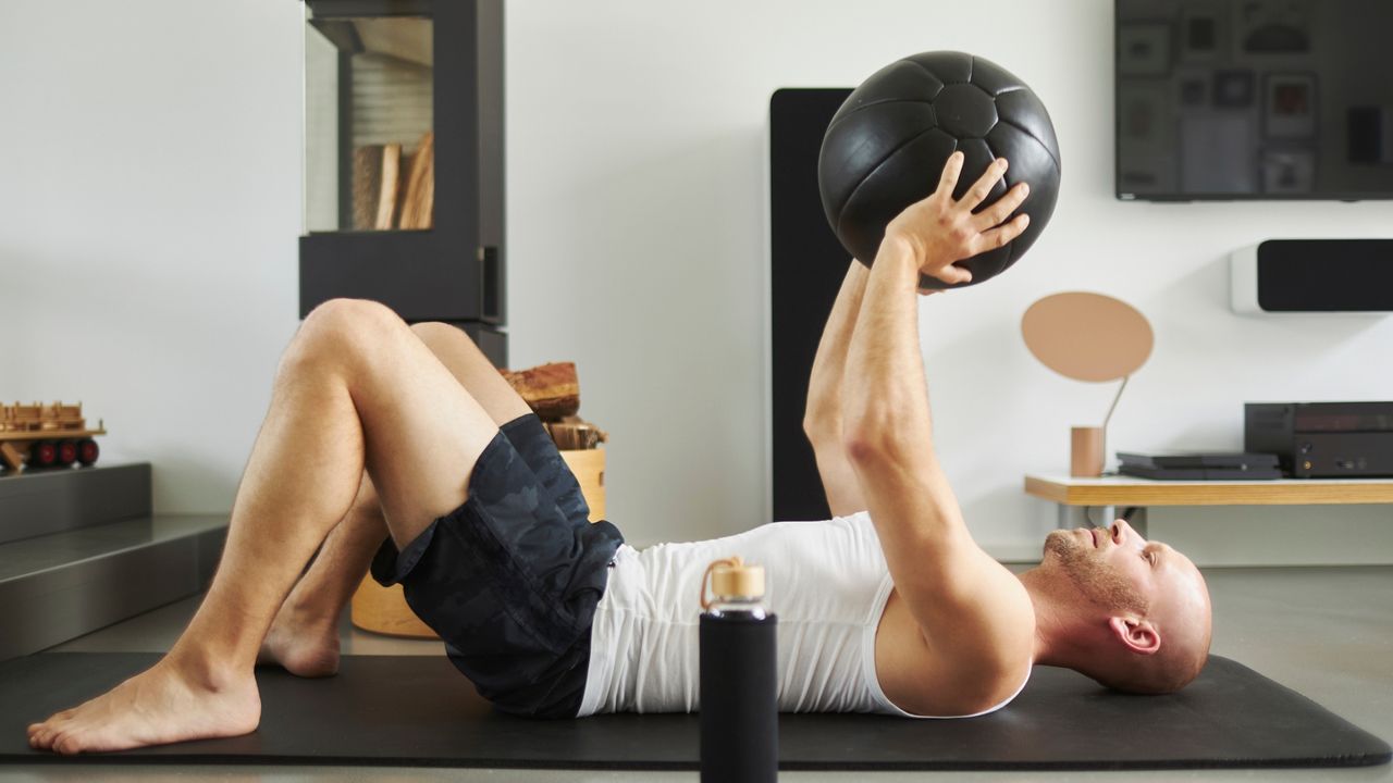 man lying on his back on a black exercise mat sideways to the camera with knees up and feet on the floor, holding a black ball overhead. there's a living room setting behind him.