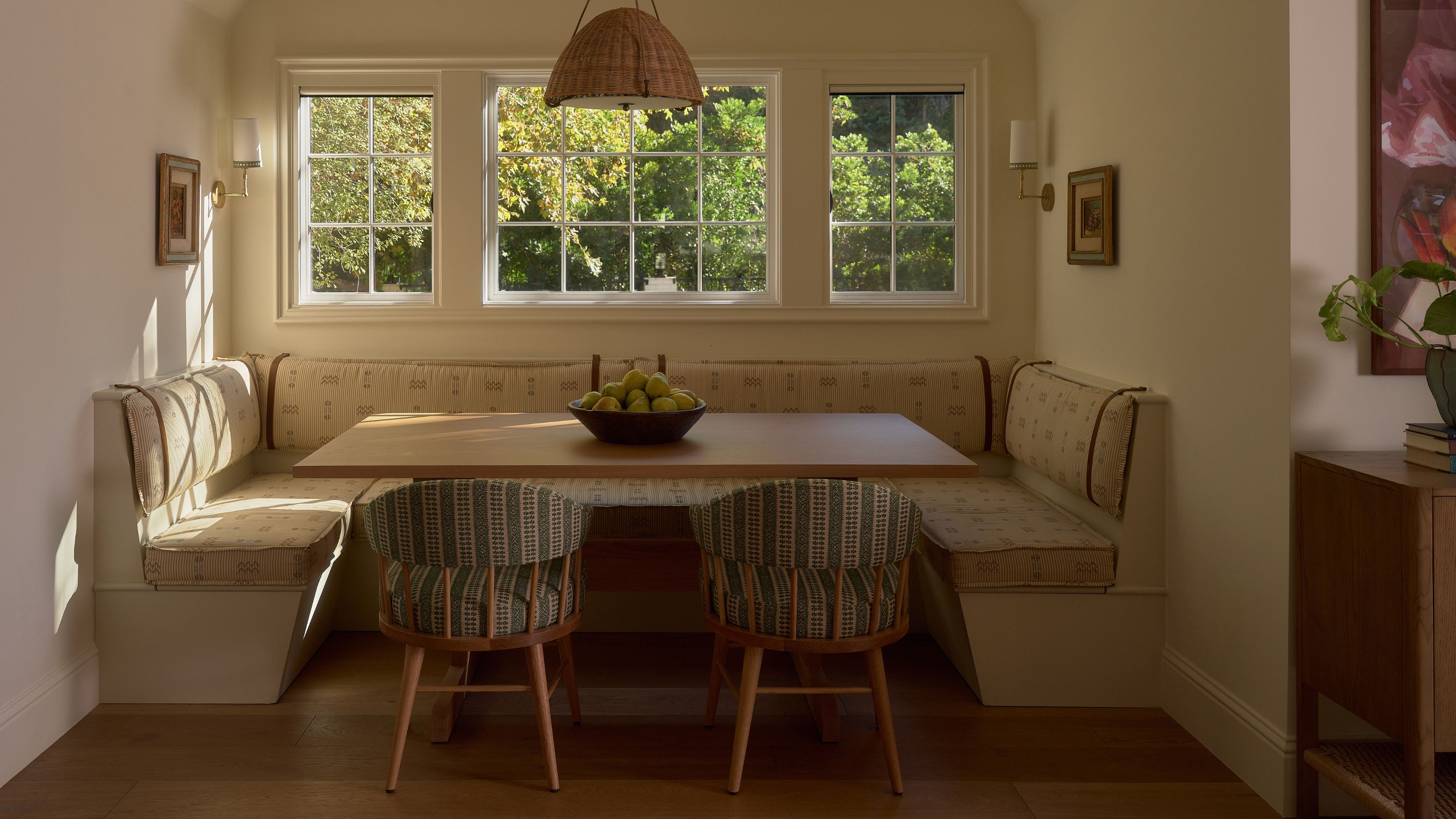 a cozy breakfast nook with a large window, banquette seat, wooden table and two print chairs with a rattan ceiling light up ahead