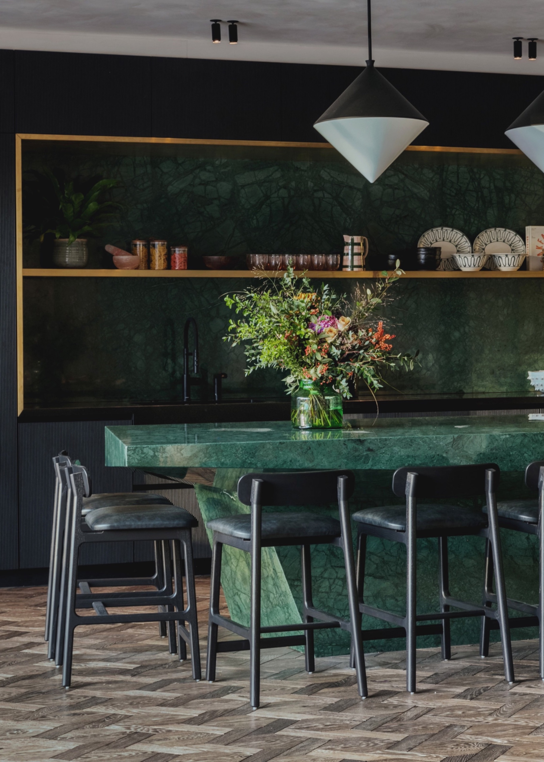 a modern kitchen with a sculptural green stone kitchen island, black bar stools, gold trim on backsplash, and vase of flowers, with two pendant lights hanging over the island