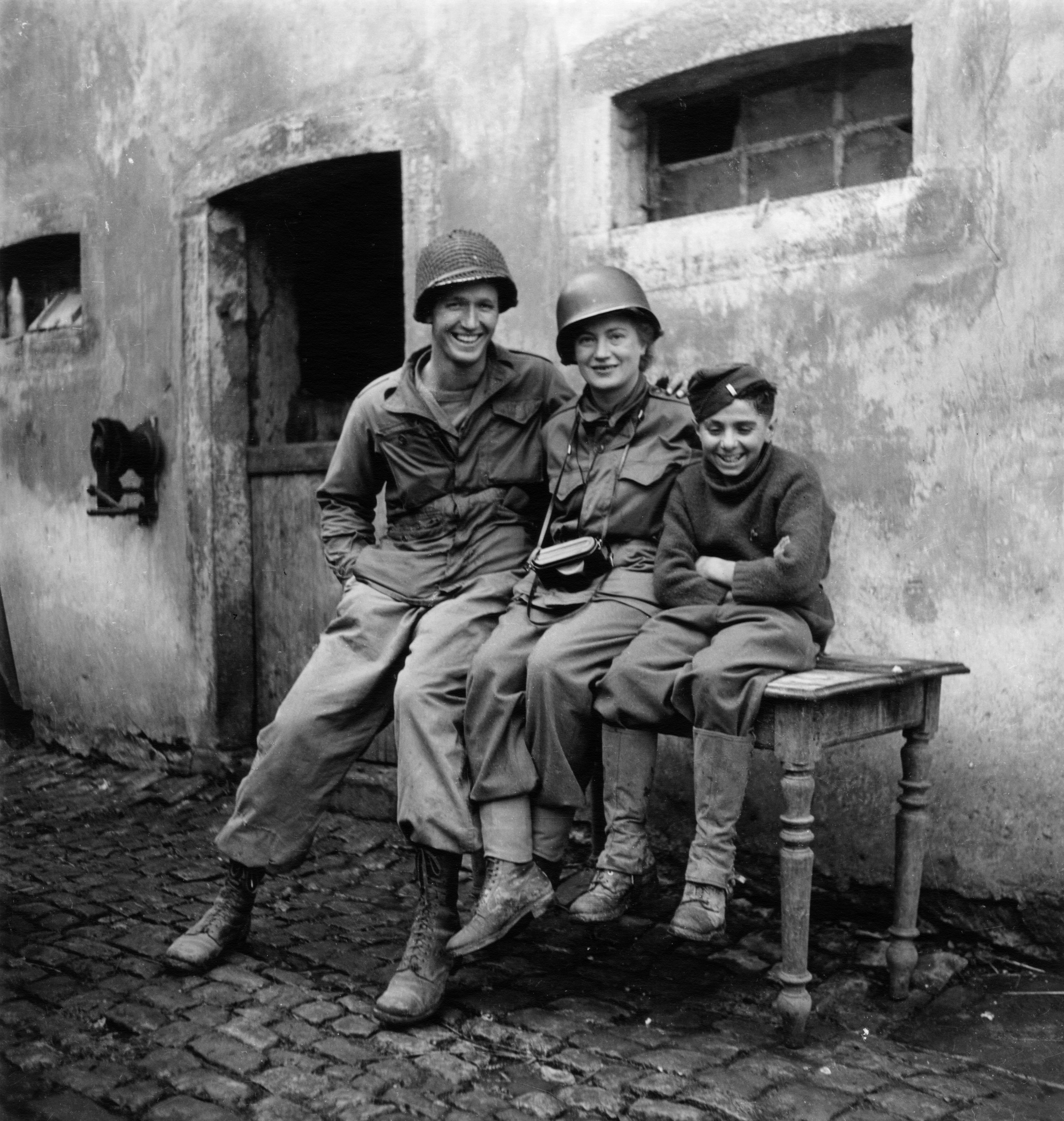 Lee Miller sits grinning between a towering American soldier and a small French boy against a cobbled Luxembourg courtyard wall, her helmet on and camera round her neck.