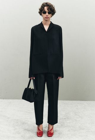 The frame bag trend is shown in the resort collection of Khaite with a model wearing a black blouse, black trousers, red pumps, and a black frame bag