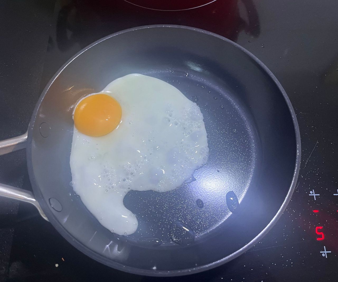 A black ceramic frying pan on a black stovetop with an egg frying inside.