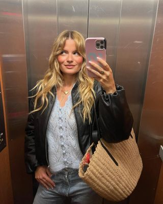 Woman standing in elevator taking mirror selfie wearing black leather jacket, light blue Sezane sweater, and wicker basket bag.