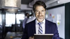 A businessman stands in his office holding a tablet.