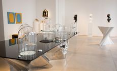 Gallery with busts on white pdiums and a glass top table with metal base with glass dome displays on top