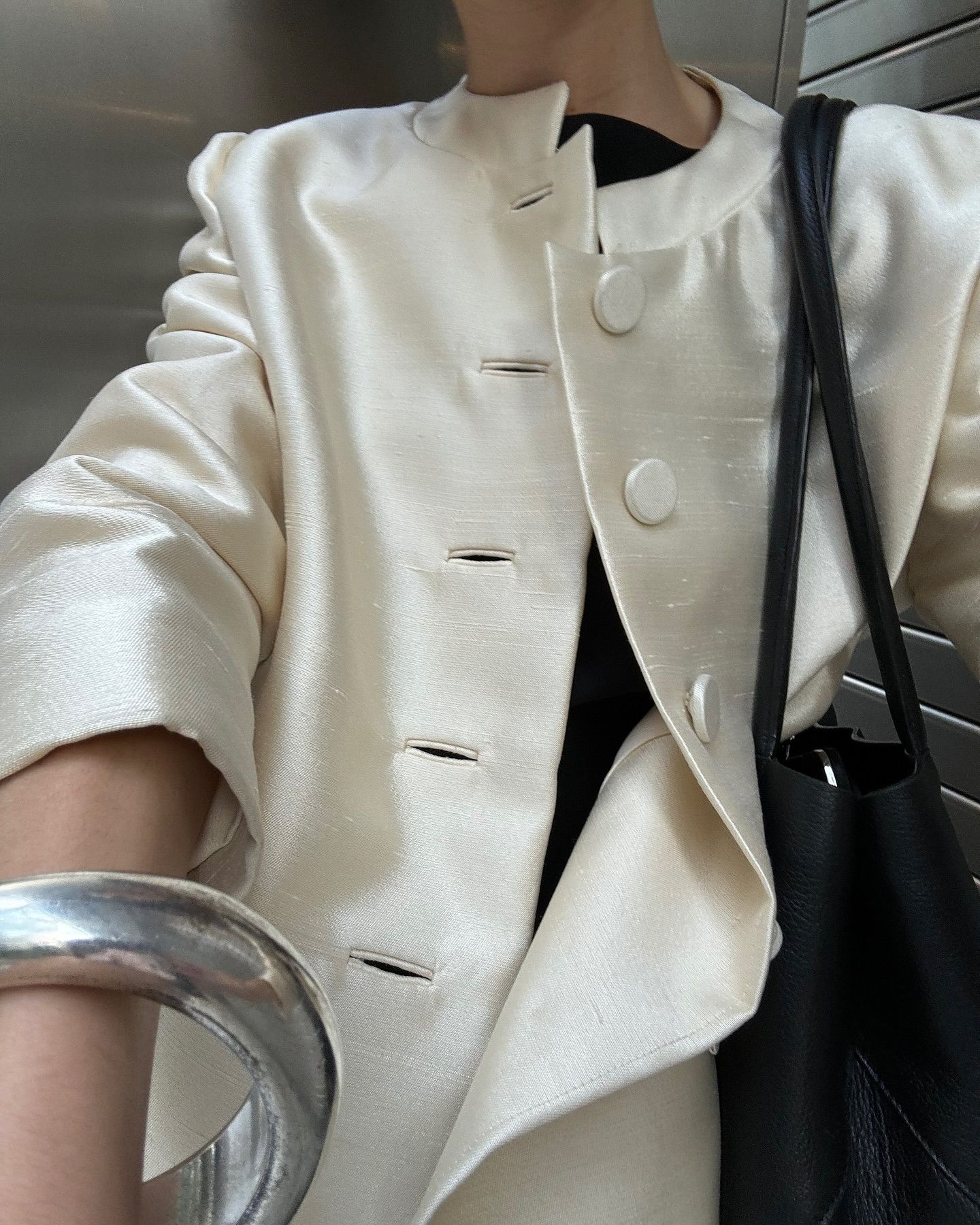 Influencer @andrea____ricci wears a cream satin high-neck blazer with a silver bangle and black bag.