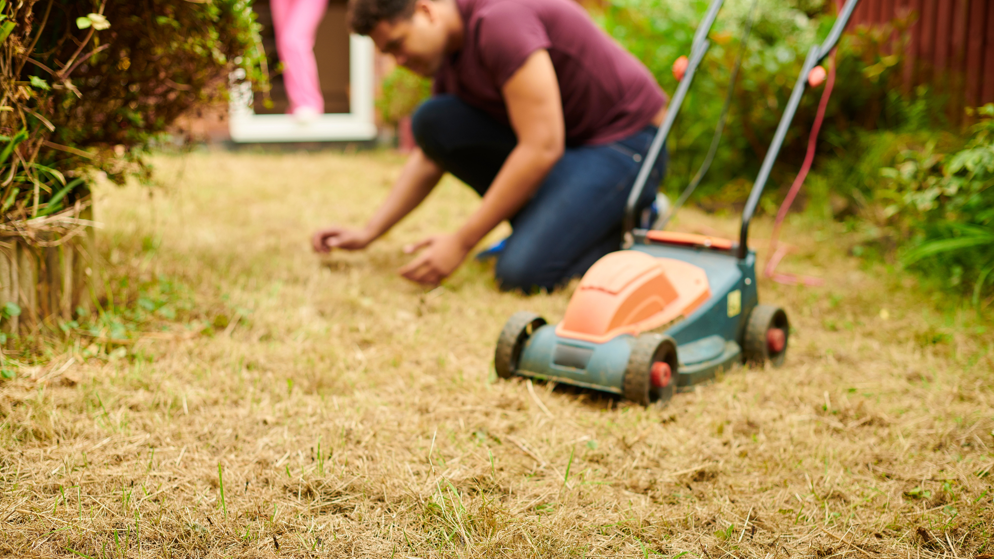 Person kneeling on dry brown grass with a lawn mower next to them