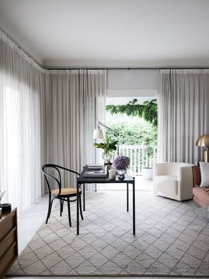 black desk and chair in living space with patterned rug, cream drapes, armchair, sofa, desk lamp, plants 