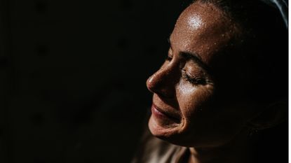 healthy smiling woman with beads of sweat on her face, lit up against dark background to represent what causes excessive sweating