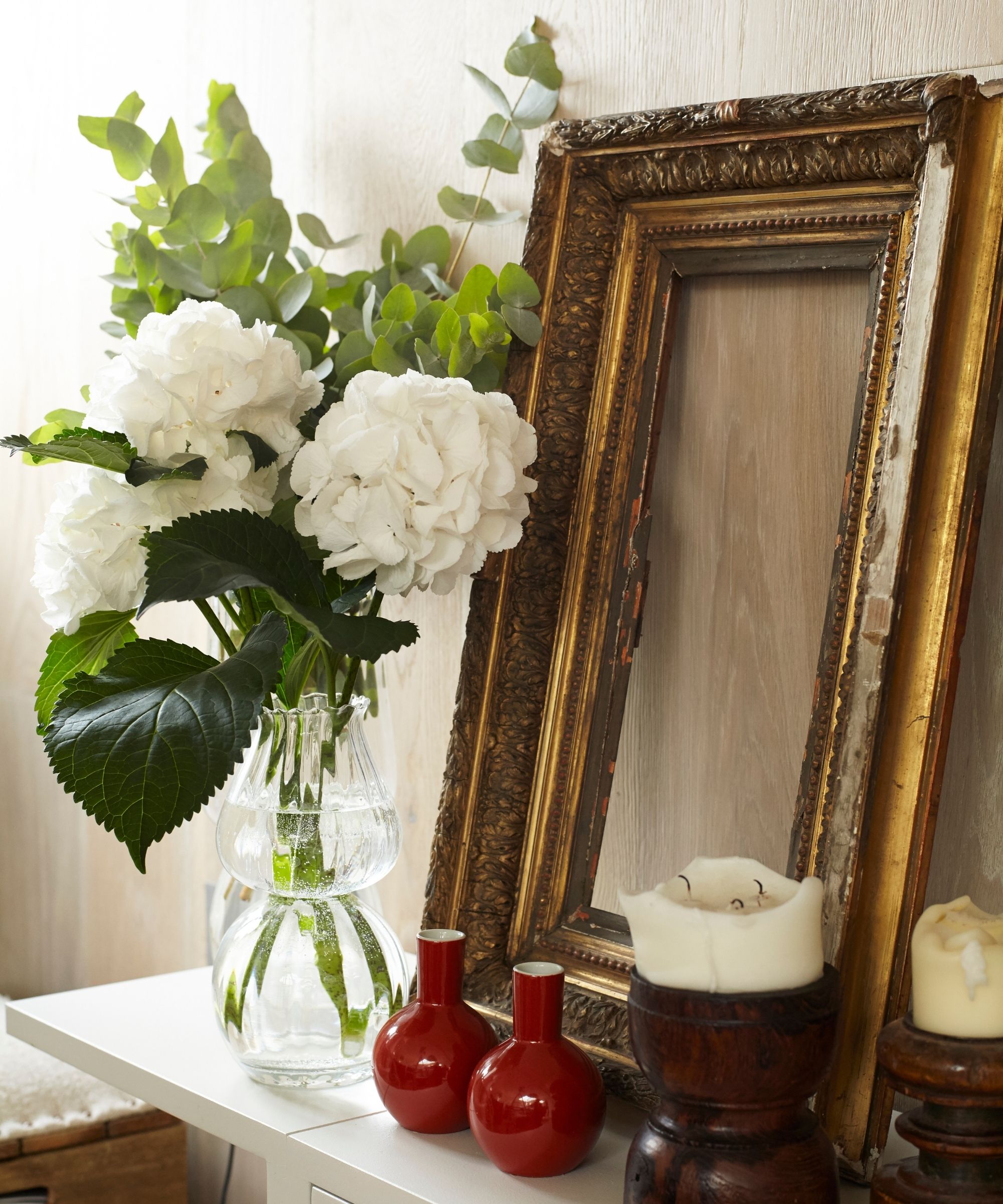 Gold frame leaning on a wooden wall on a white mantelpiece with white hydrangeas in a glass vase, red mini vases, and two white pillar candles in polished wood holders.