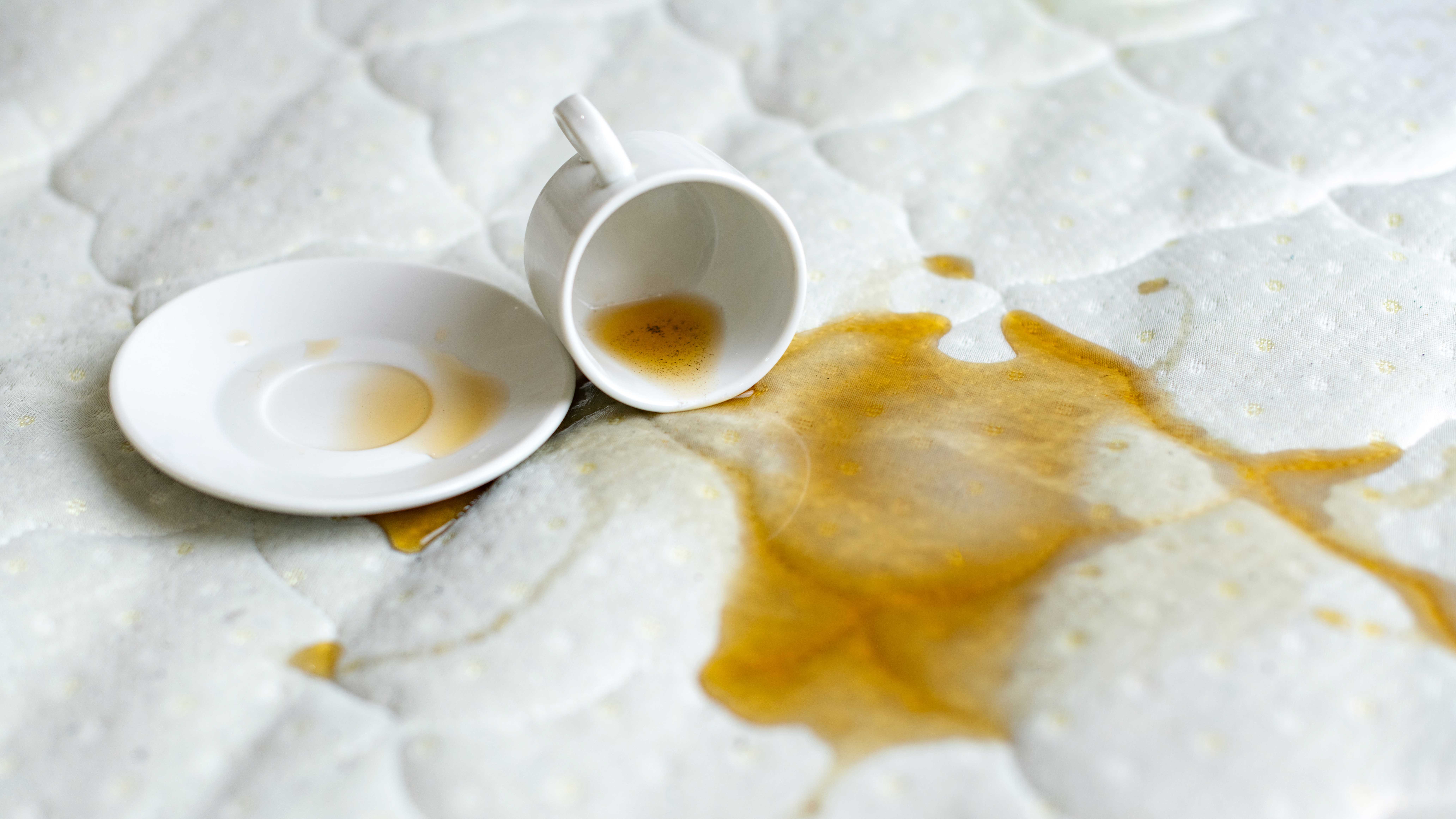 Изображение, показывающее, как чашка черного чая разливается по белому матрасу.