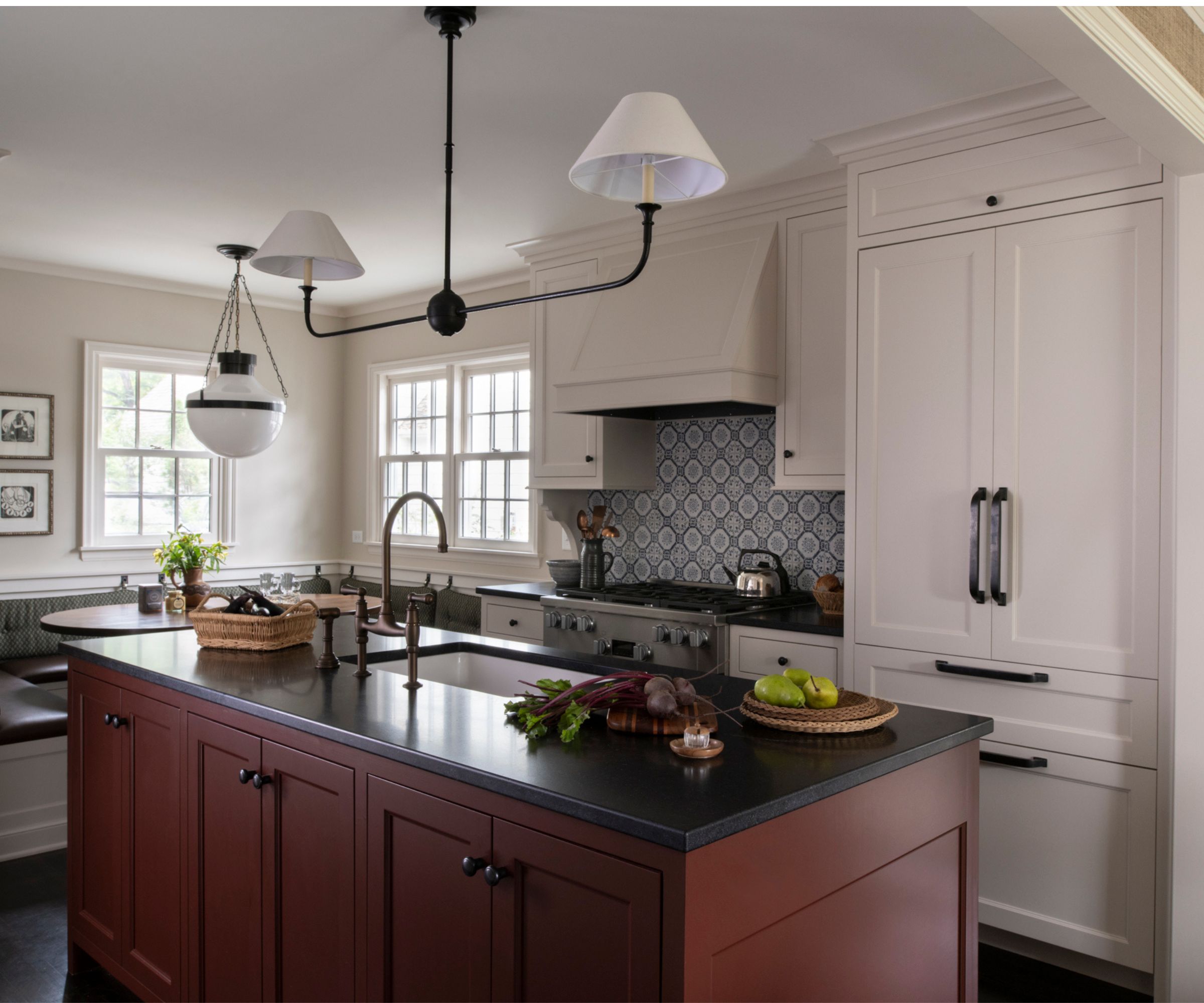 white farmhouse style kitchen with a blue patterned tile backsplash, red kitchen island with dark countertops, breakfast nook in the corner beneath the windows and white and black vintage style lighting