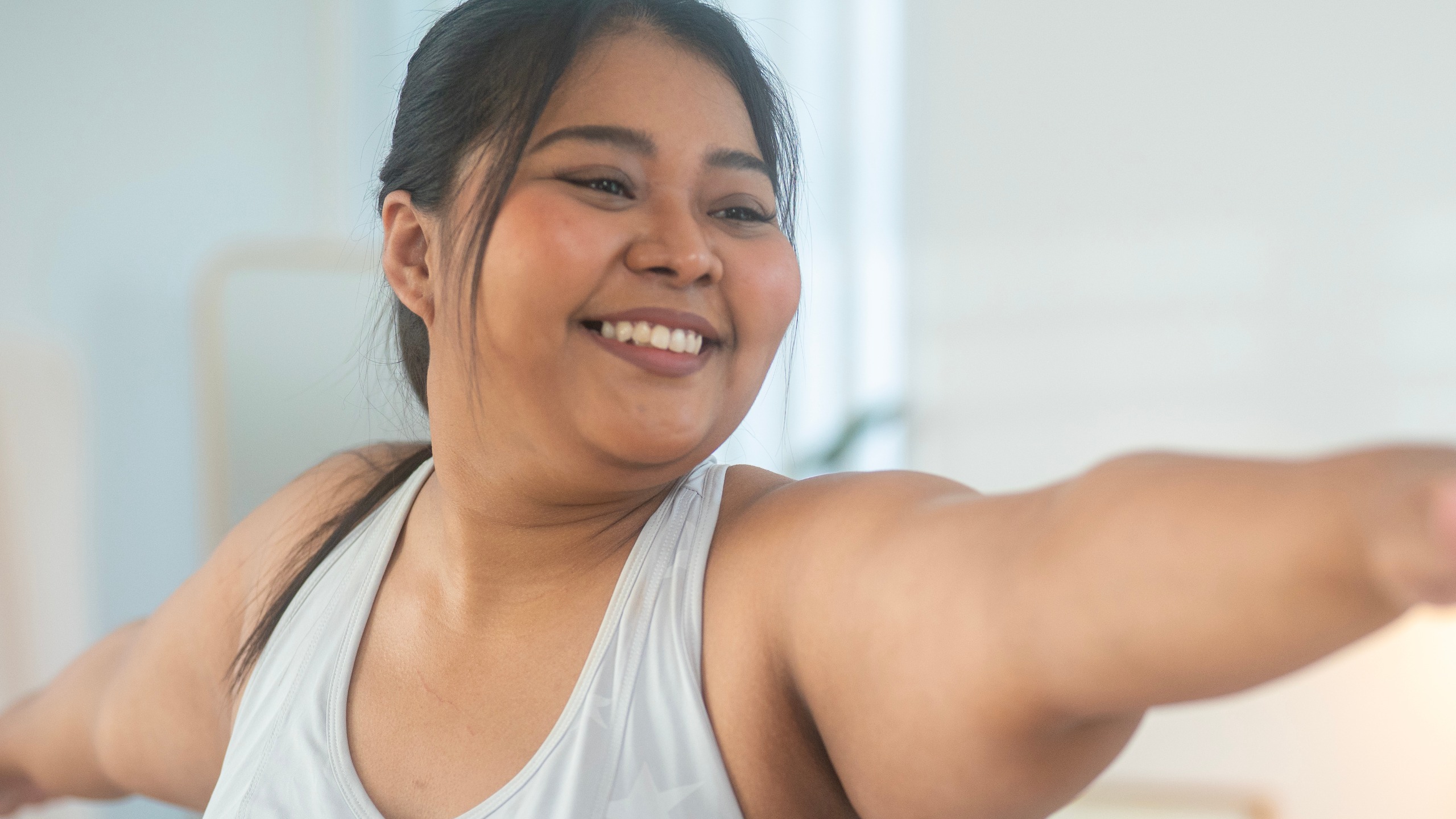 A woman performs holds warrior two yoga pose while smiling. Her arms are outstretched at shoulder height. 