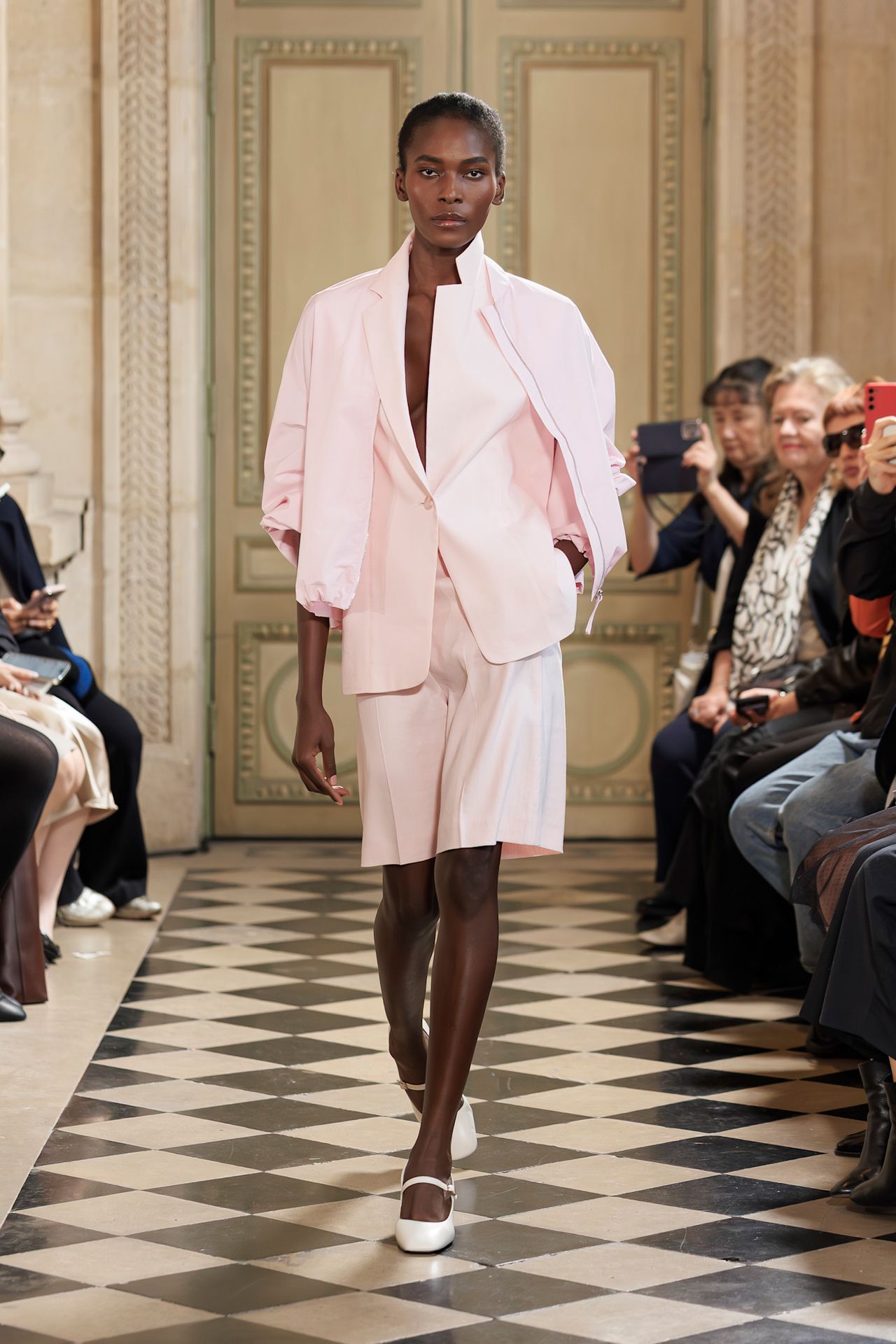 A Laur&egrave;l model wearing a pale pink jacket layered on top of a matching blazer and pleated midi-shorts and a pair of white mary jane ballet flats.