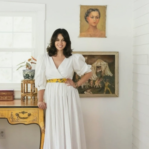 Image of a woman in a white dress standing in a white room with an antique yellow console table beside her and antique art on the walls.