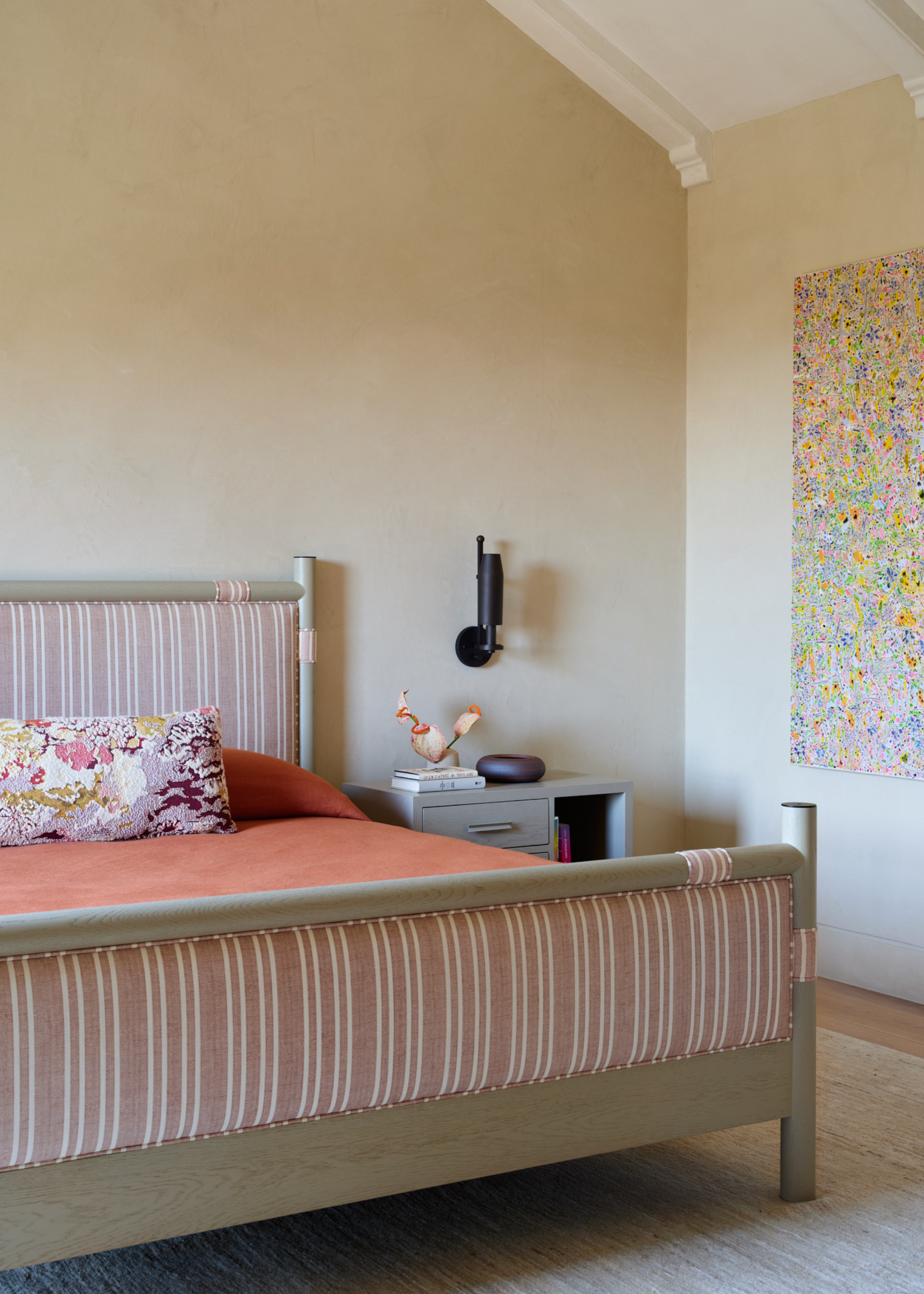 A cream bedroom with a wood bed featuring a striped upholstered foot and head with a patterned pillow and a coral duvet beside a side table, a black wall sconce, and a colorful painting