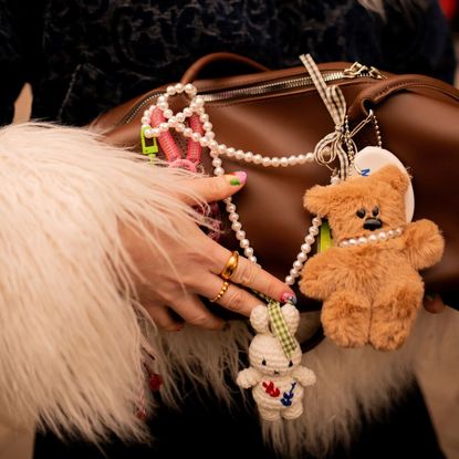 a woman's hand holding a brown handbag with various charms and decorations including a teddy bear charm and a bunny charm with some faux pearls. 