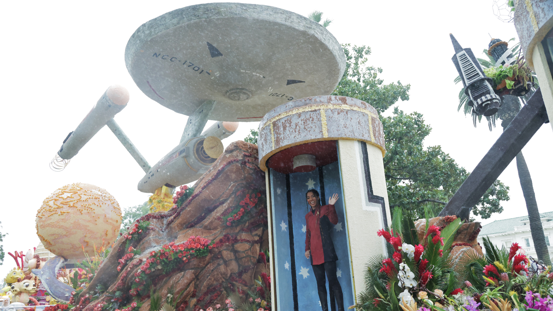 A woman in a red shirt and black pants stands in a tall open cylinder with a blue and white dotted pattern with a large spaceship above the cylinder.