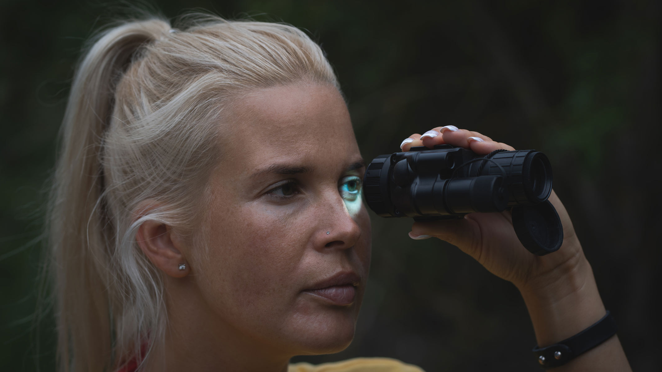 A blonde woman looking through a night vision monocular.