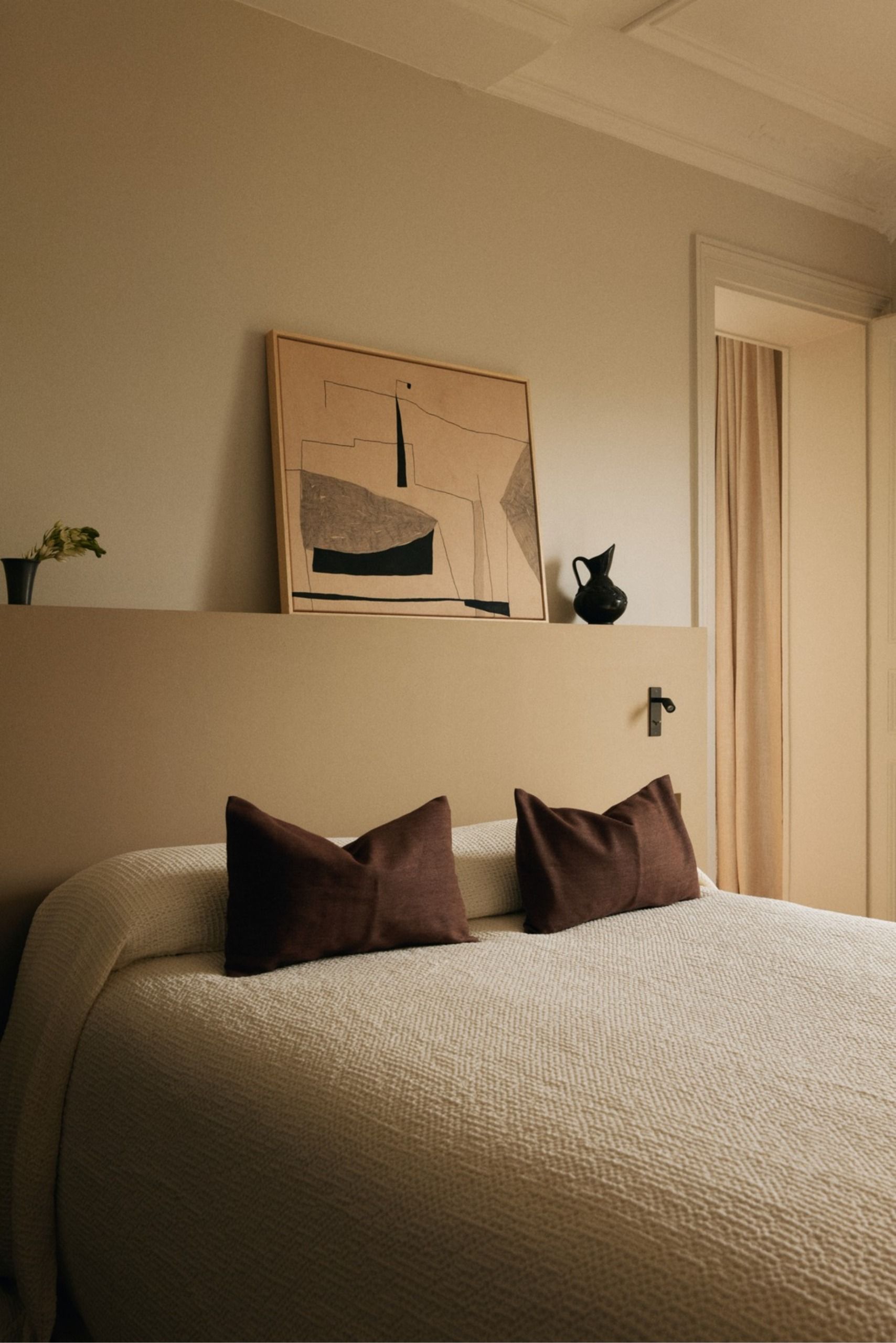 Neutral bedroom with a textured cream bedspread, dark brown cushions, abstract artwork, a beige half-height wall and a black ceramic vase