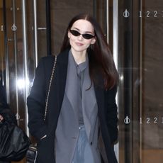 Dove Cameron wearing a black coat, grey top, gray pants, and black heels